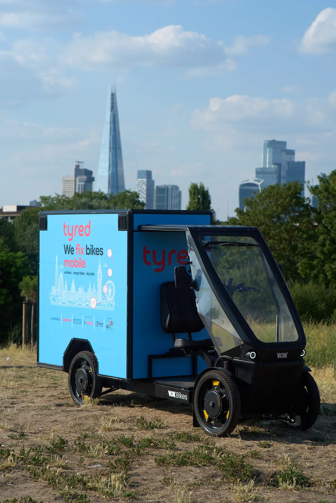 A Tyred-branded cargo bike parked in a London park with The Shard and the City skyline behind.