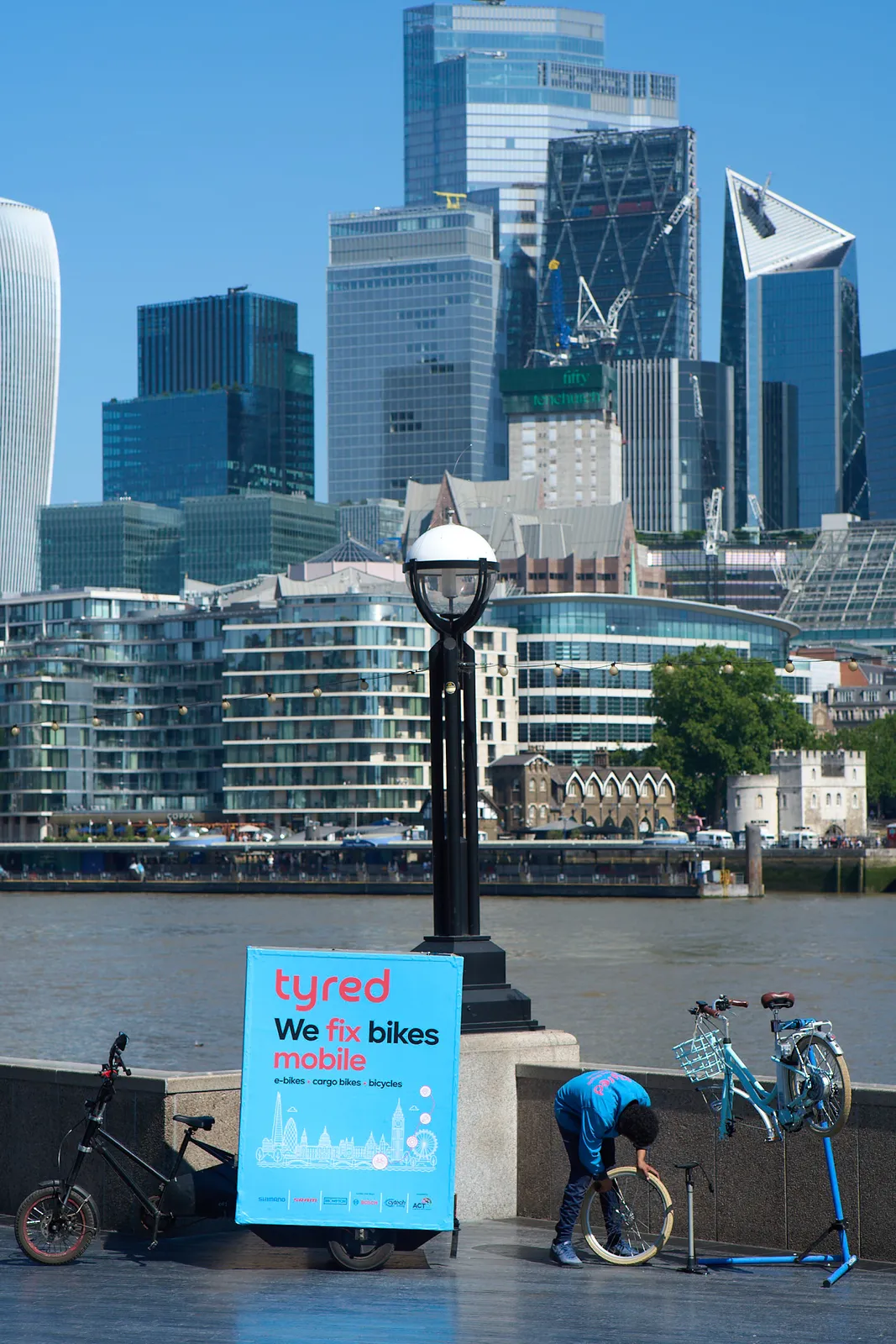 A Tyred mechanic inflating a bike tyre beside the Thames, with the City of London skyline behind.