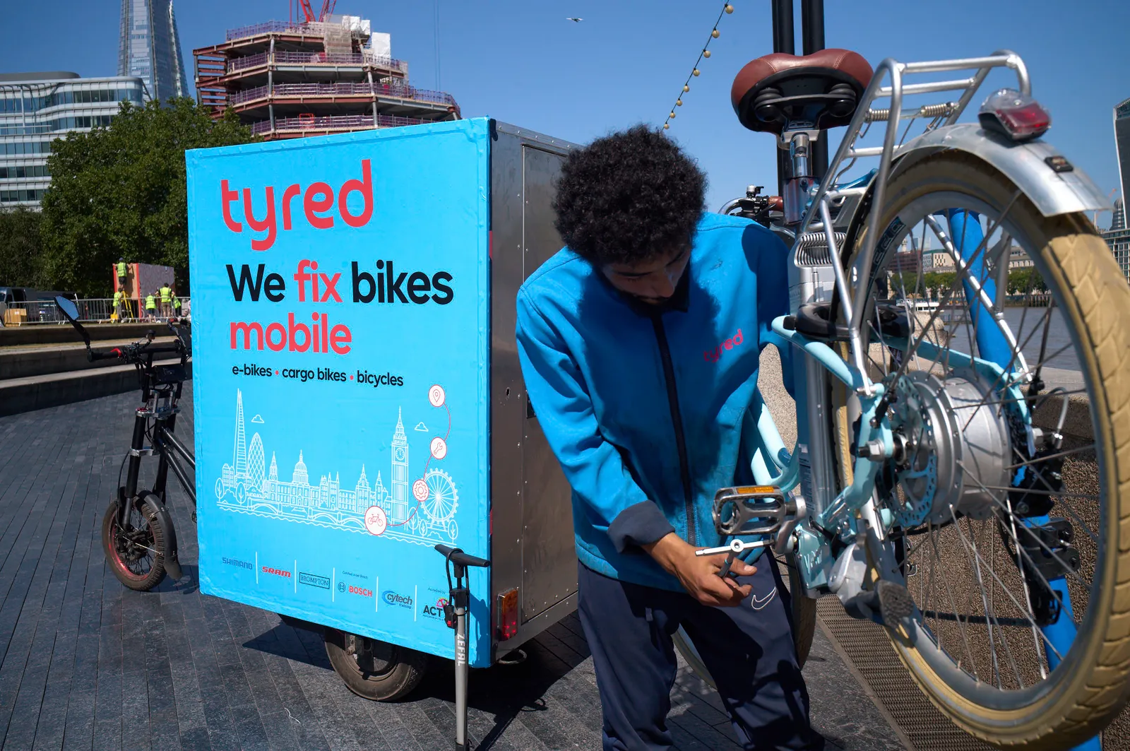 A Tyred mechanic in a branded jacket repairing a bike's rear wheel beside the mobile service trailer in London.