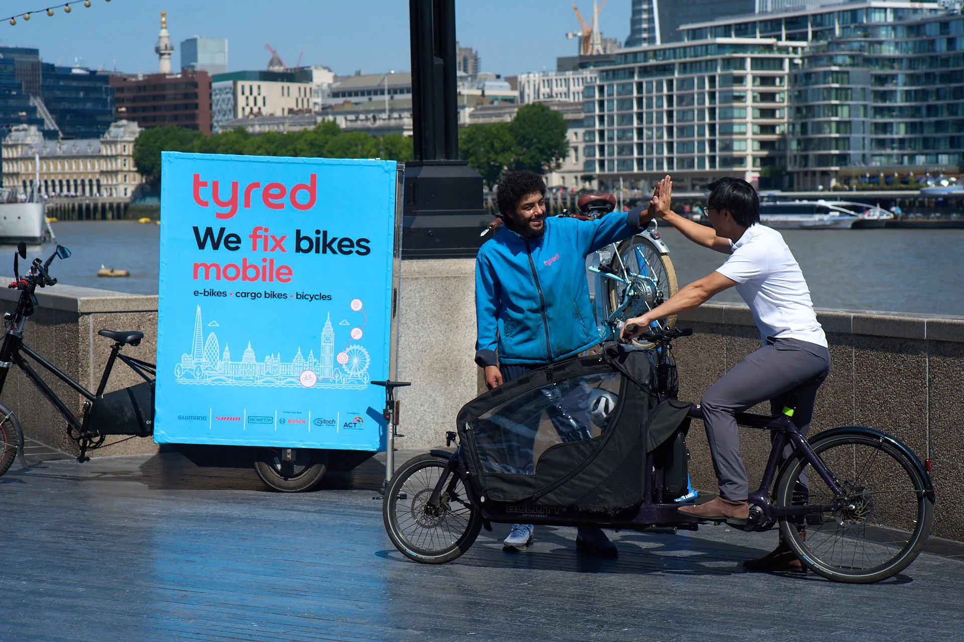 A Tyred mechanic high-fiving a cargo bike customer after a riverside repair in London.