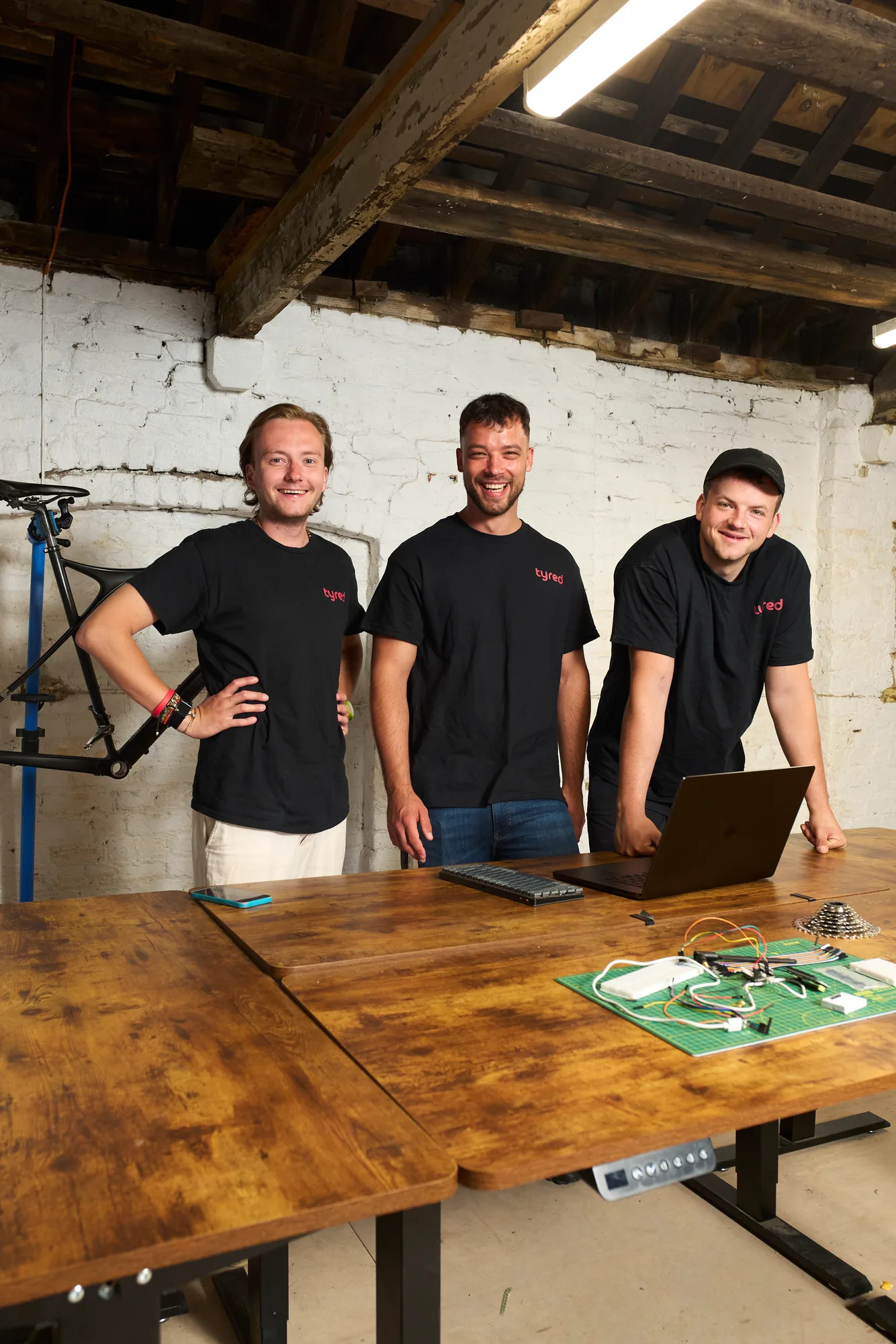 The Tyred founding team in their workshop, with a bike on a repair stand behind them.